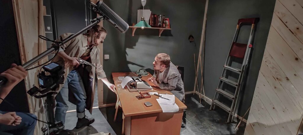 Viiksekäs mies osoittaa aseella kirjoituspöydän takan istuvaa henkilöä 
 Elokuvan lavasteissa. Etualalla näkyy järjestelmäkamera ja mikrofoni.